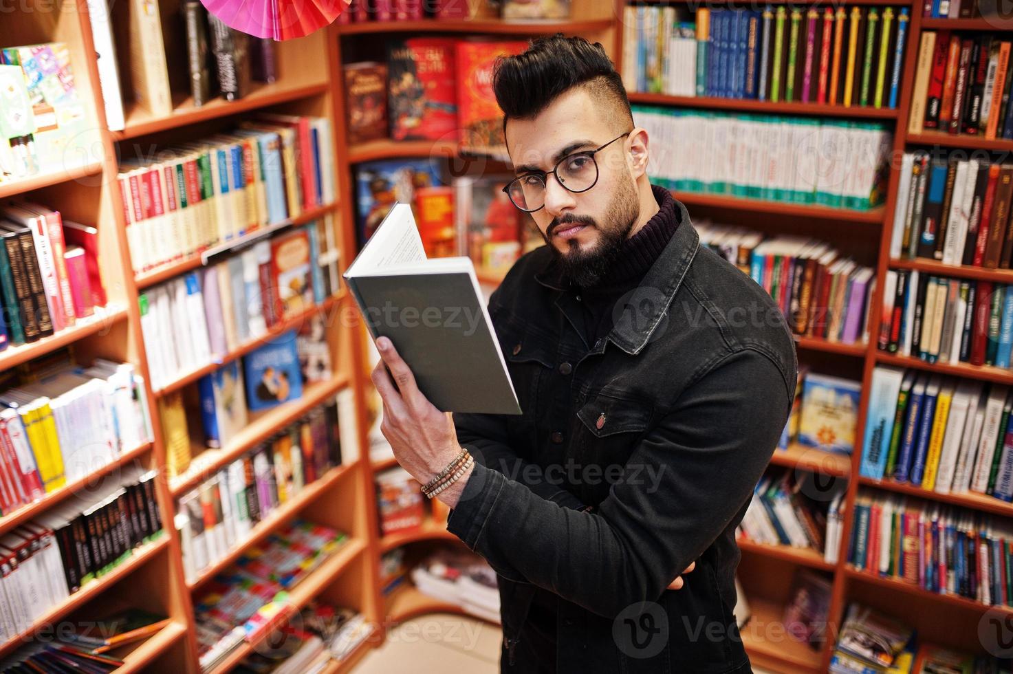 estudiante árabe alto e inteligente, vestido con chaqueta negra de jeans y anteojos, en la biblioteca con un libro en las manos. foto