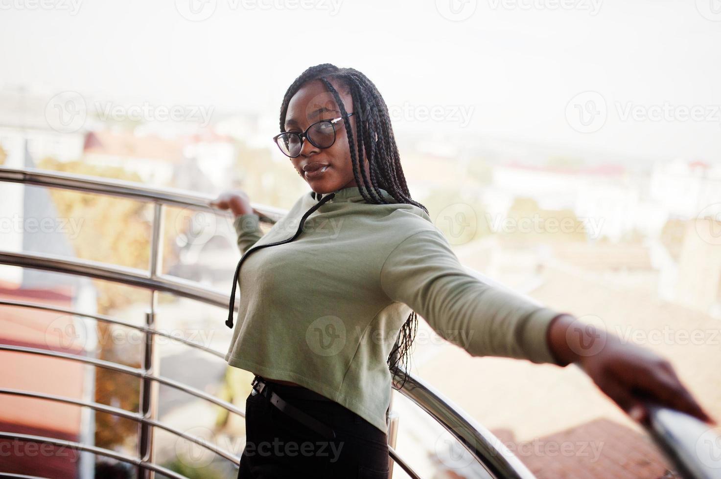 City portrait of positive young dark skinned female wearing green hoody and eyeglasses standing at balkony. photo