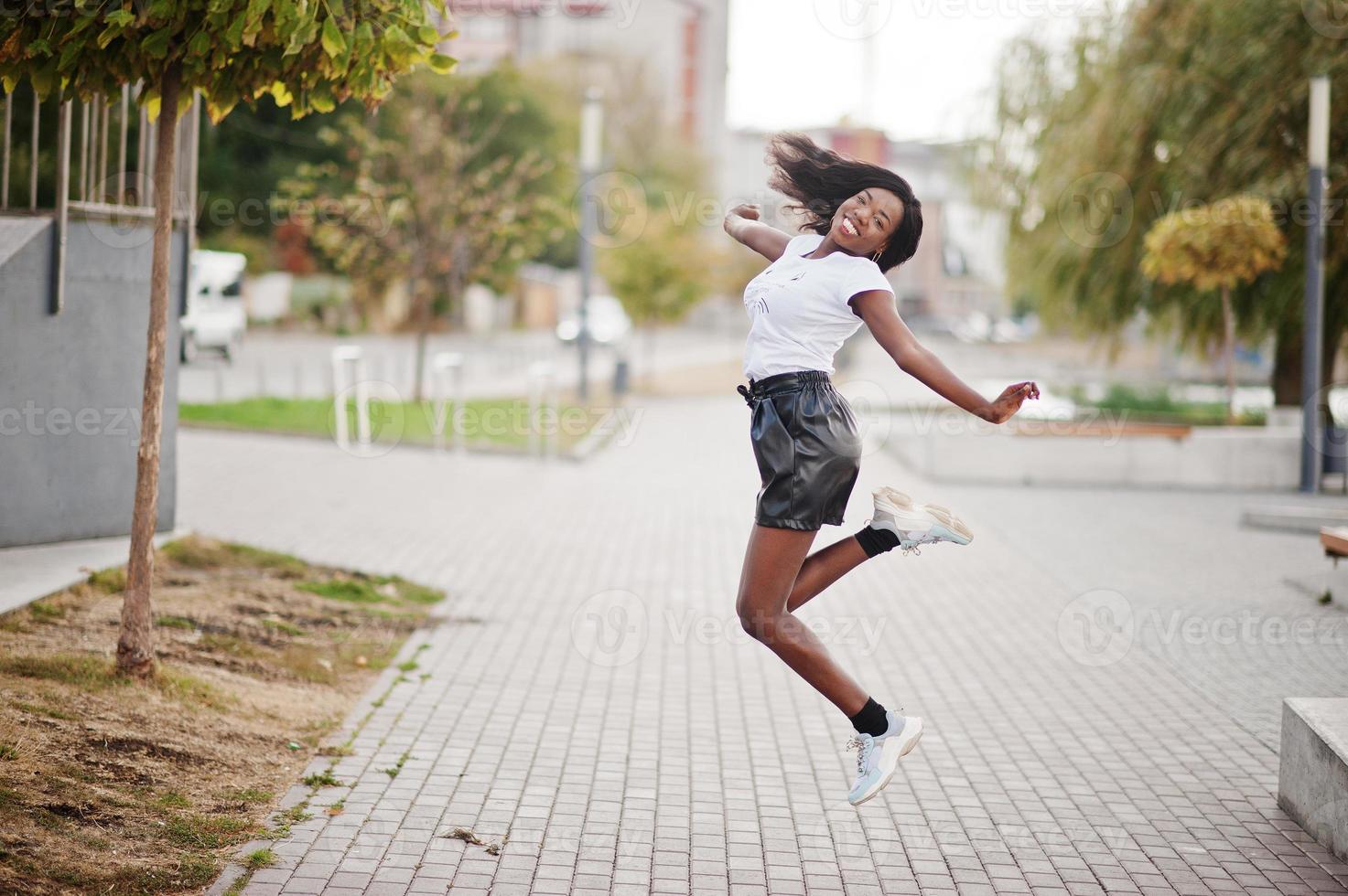 African american dark skinned slim model posed in a black leather shorts and white t-shirt. She jumping at the air. photo