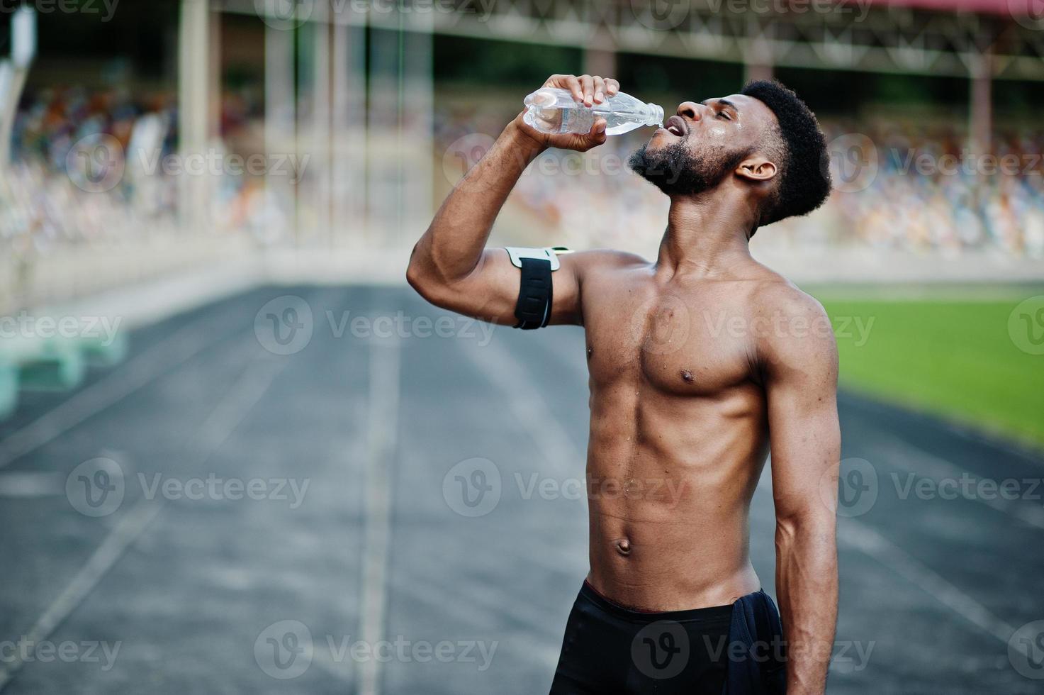 atleta masculino afroamericano sexy deporte torso desnudo hombre con brazo deportivo corriendo funda para teléfono móvil, posó en el estadio y bebiendo agua de la botella. foto