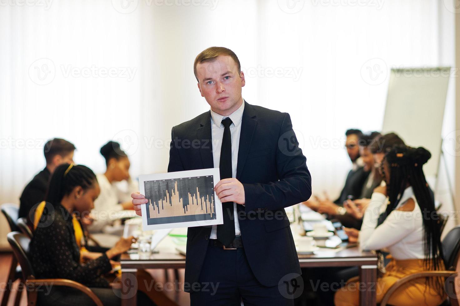 Face of handsome european business man, holding paper with diagram on the background of business peoples multiracial team meeting, sitting in office table. photo