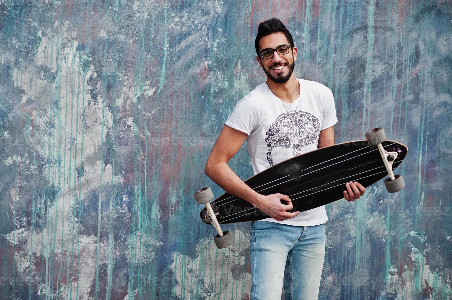 hombre árabe de estilo callejero con anteojos con longboard posado contra una pared de color, como si tocara la guitarra. foto