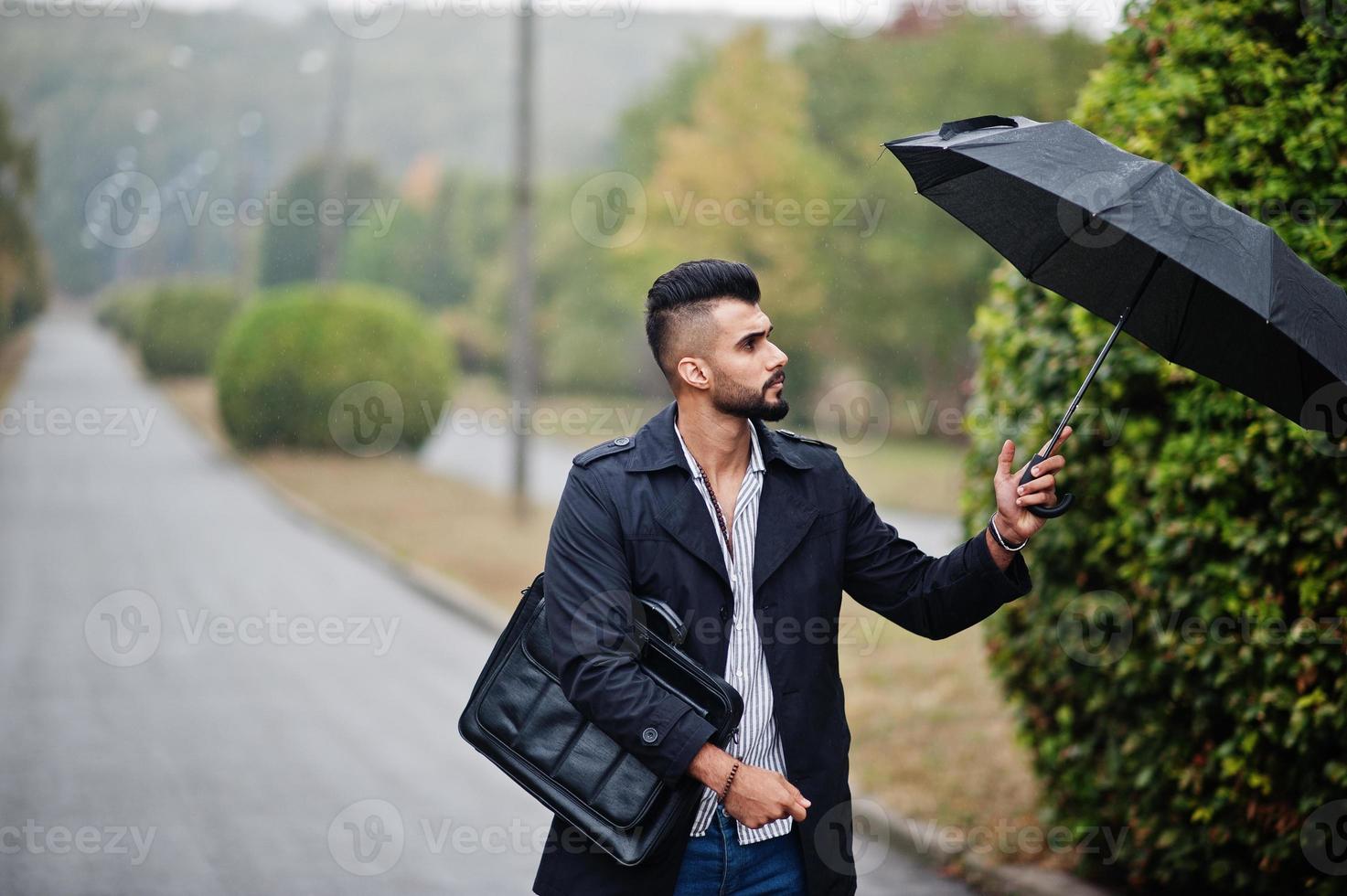 Fashionable tall arab beard man wear on black coat with umbrella and bag case posed at rain weather day. photo