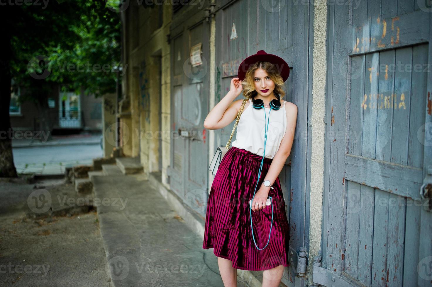 Fashionable and beautiful blonde model girl in stylish red velvet velour skirt, white blouse and hat, posed with phone and earphones. photo