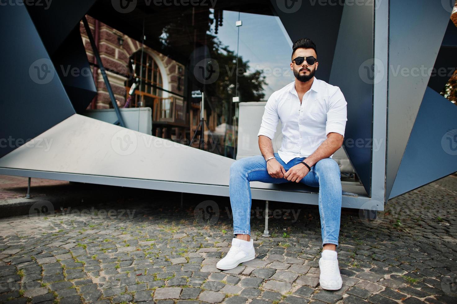 Stylish tall arabian man model in white shirt, jeans and sunglasses sitting at street of city. Beard attractive arab guy. photo