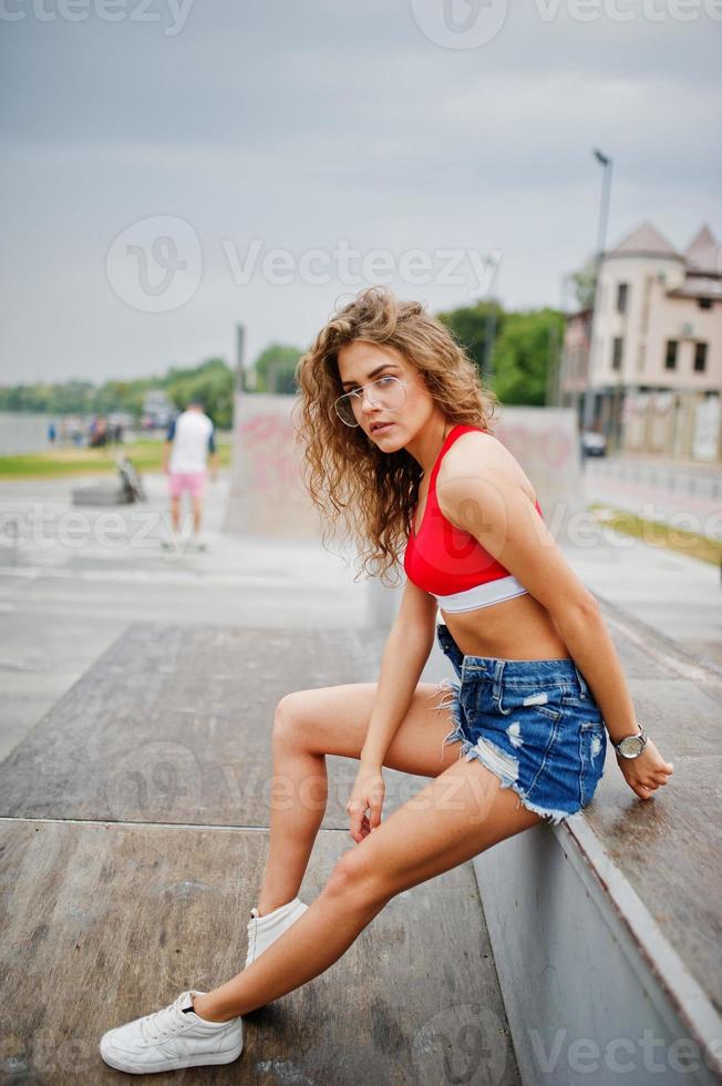 chica sexy modelo rizada en top rojo, pantalones cortos de mezclilla, anteojos y zapatillas posadas en skatepark. foto