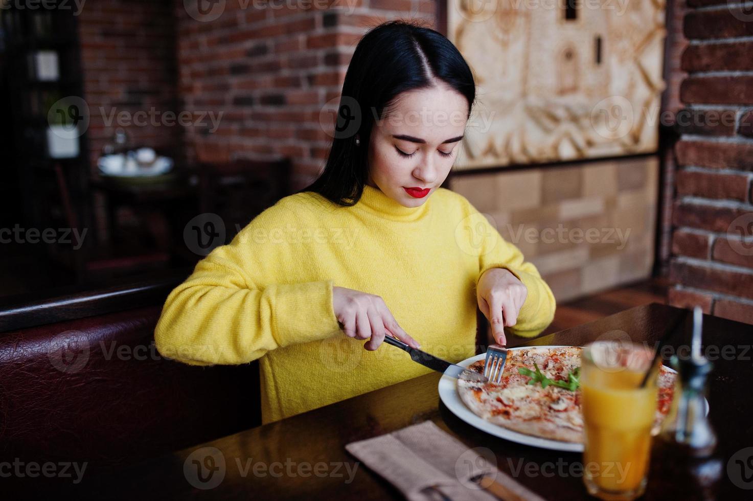 Funny brunette girl in yellow sweater eating pizza at restaurant. photo