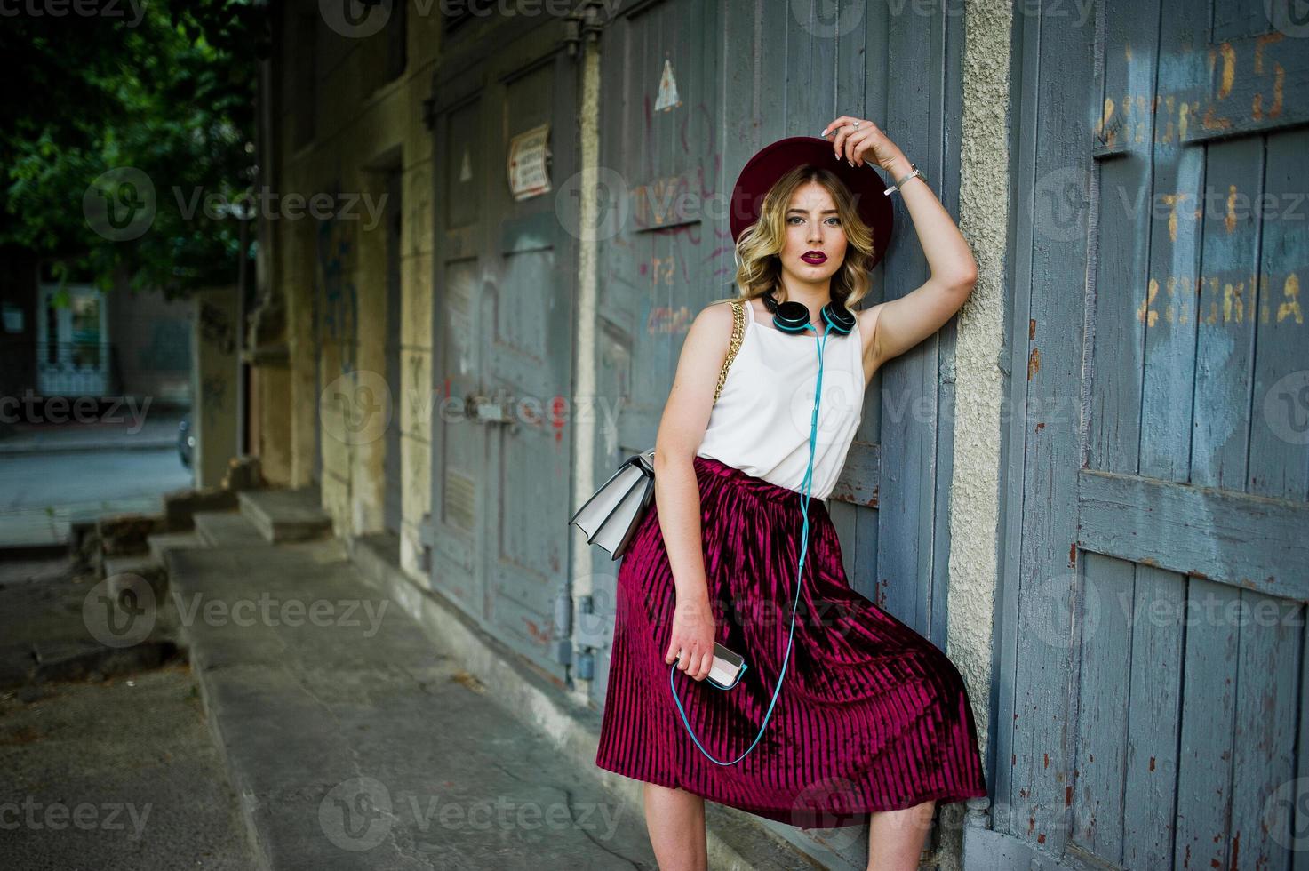 Fashionable and beautiful blonde model girl in stylish red velvet velour skirt, white blouse and hat, posed with phone and earphones. photo
