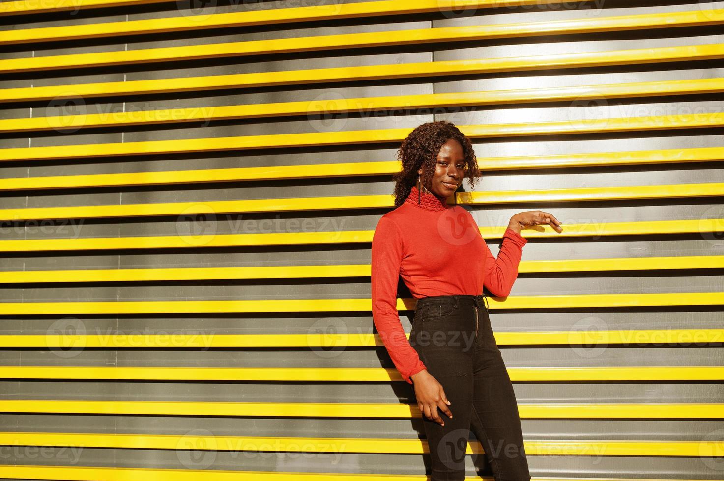 Portrait of a curly haired african woman wearing fashionable red turtleneck posing against yellow modern lines wall. photo