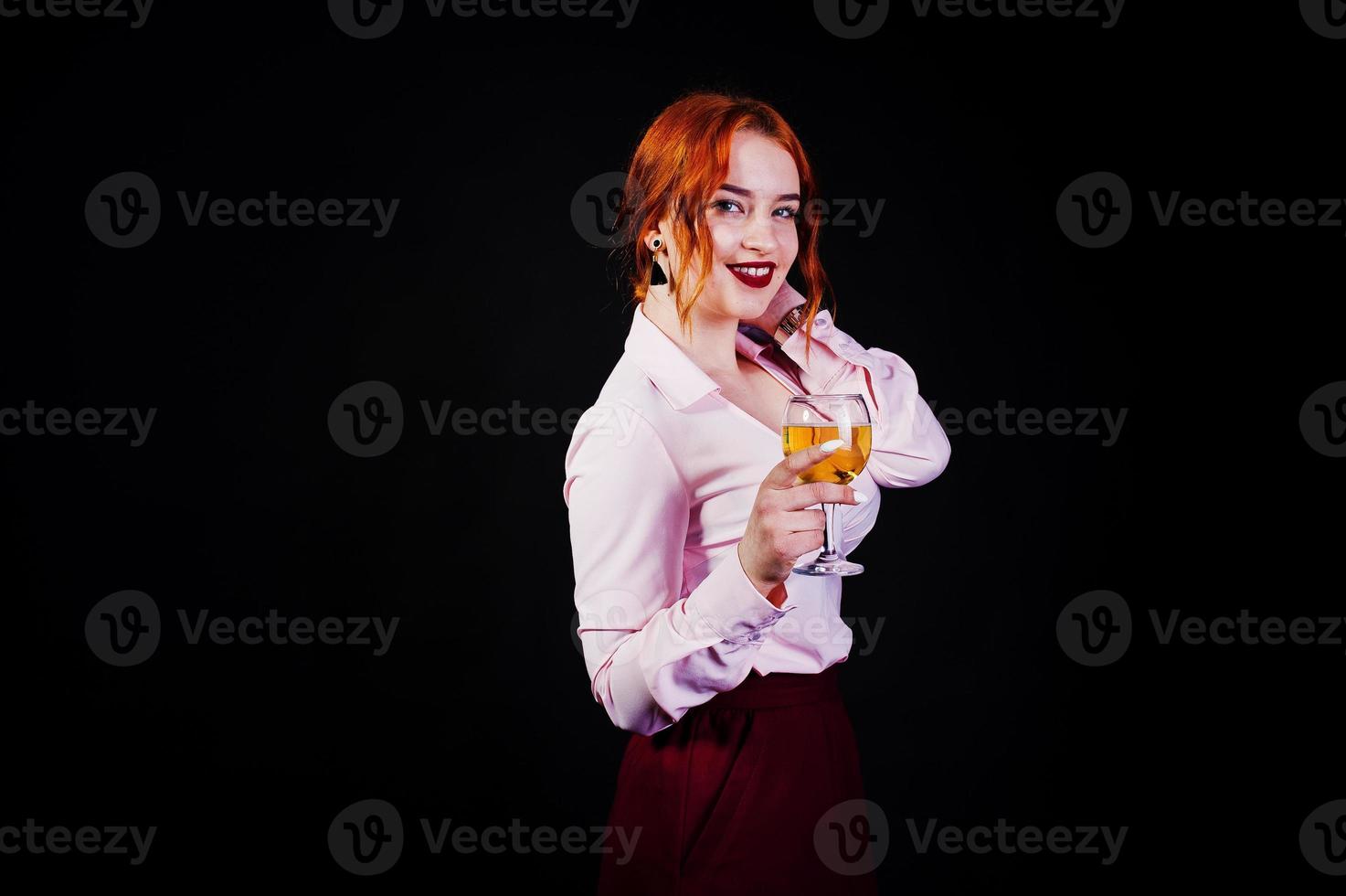 Gorgeous red haired girl in pink blouse and red skirt with glass of wine at hand isolated on black. photo
