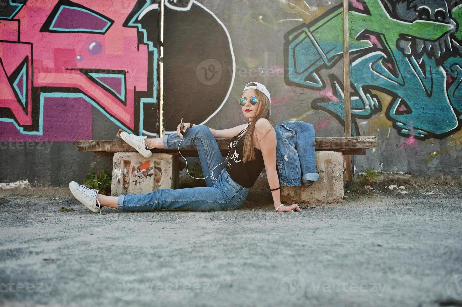 Stylish casual hipster girl in cap, sunglasses and jeans wear, listening music from headphones of mobile phone against large graffiti wall with bomb. photo