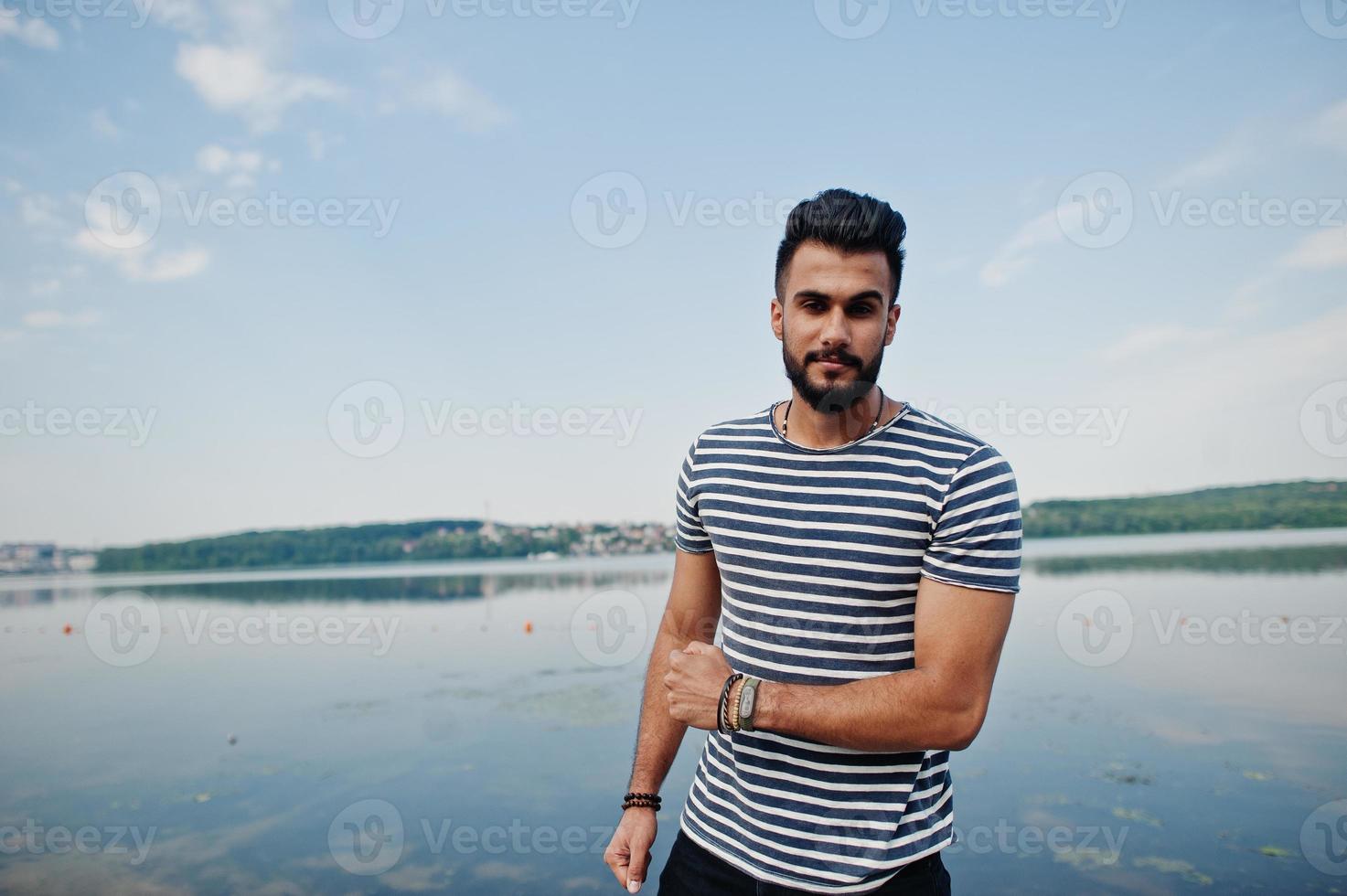 Handsome tall arabian beard man model at stripped shirt posed outdoor against lake and sky. Fashionable arab guy. photo