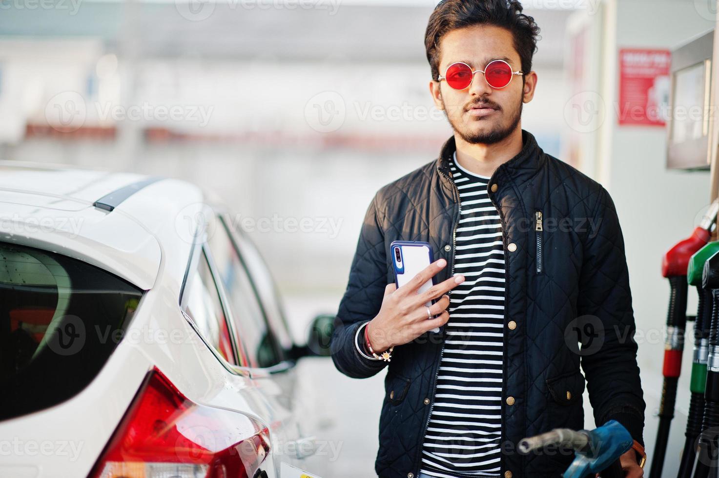 South asian man or indian male refueling his white car on gas station with mobile phone at hand. photo