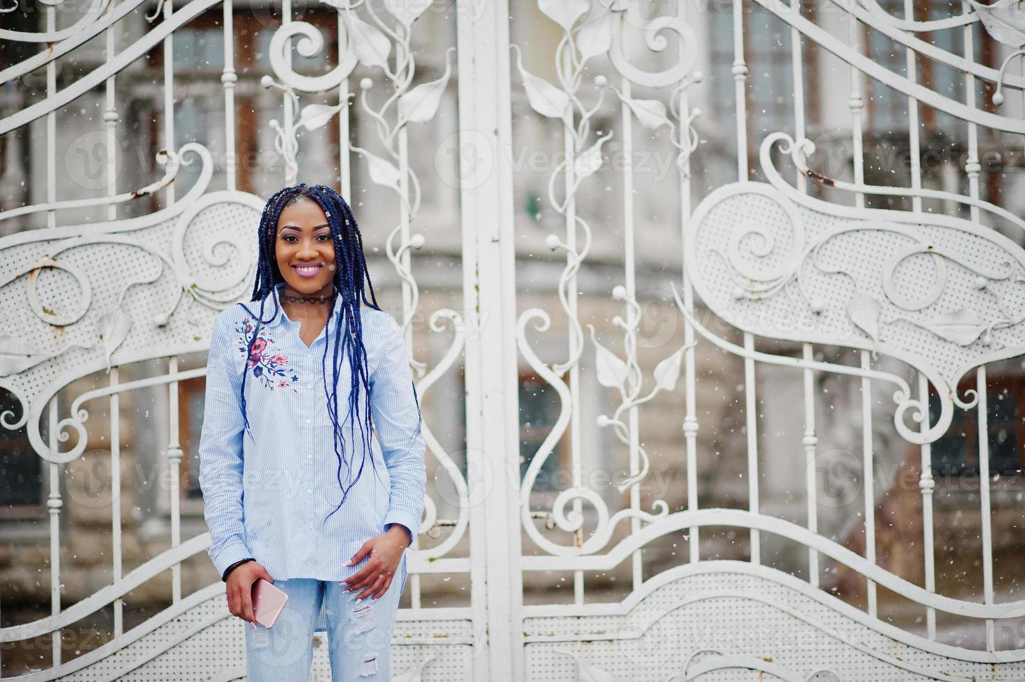 Stylish african american girl with dreads holding mobile phone at hand, outdoor against white royal gates at snowy weather. photo