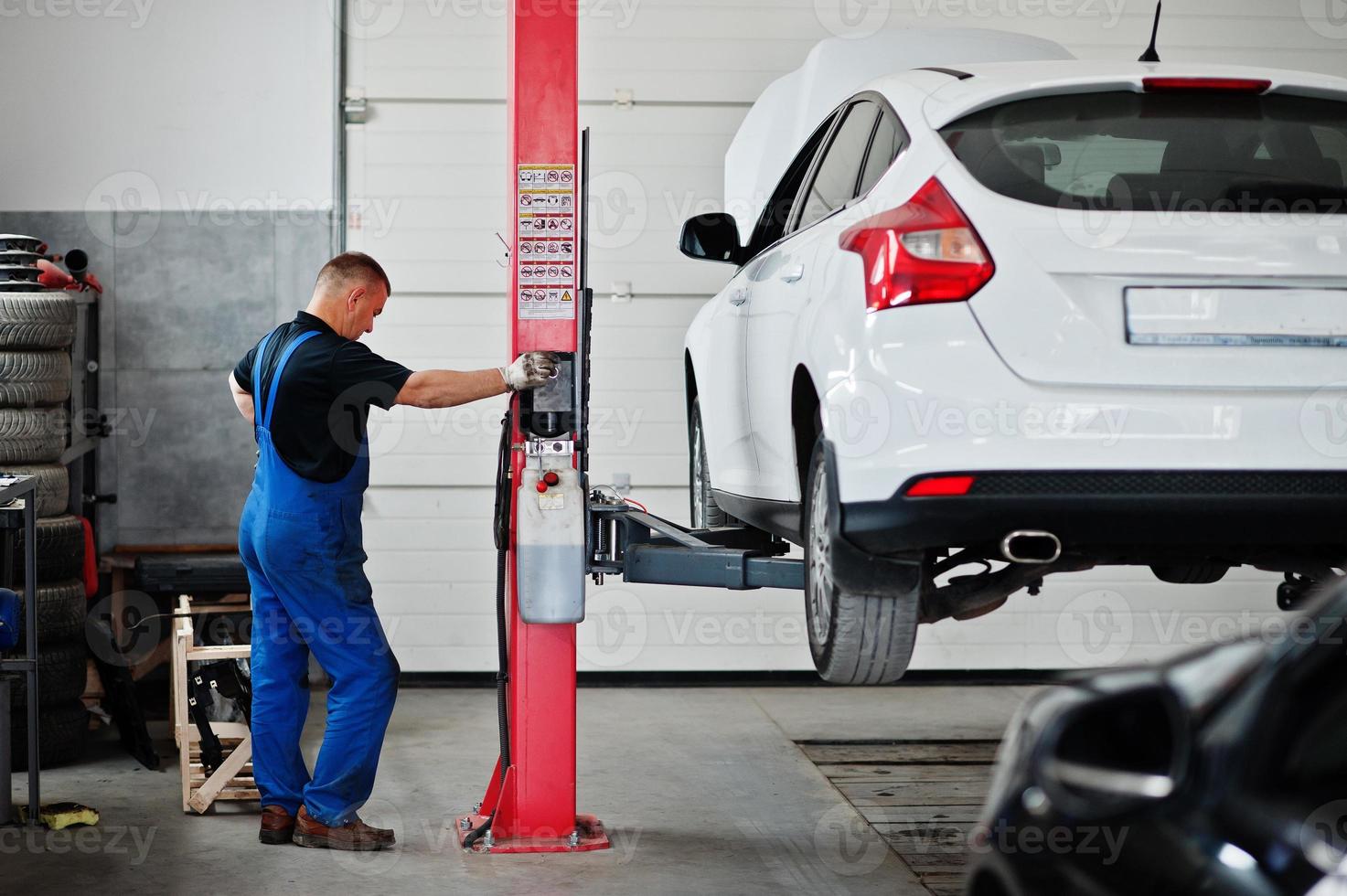 Car repair and maintenance theme. Mechanic in uniform working in auto service, press button to lifting auto. photo