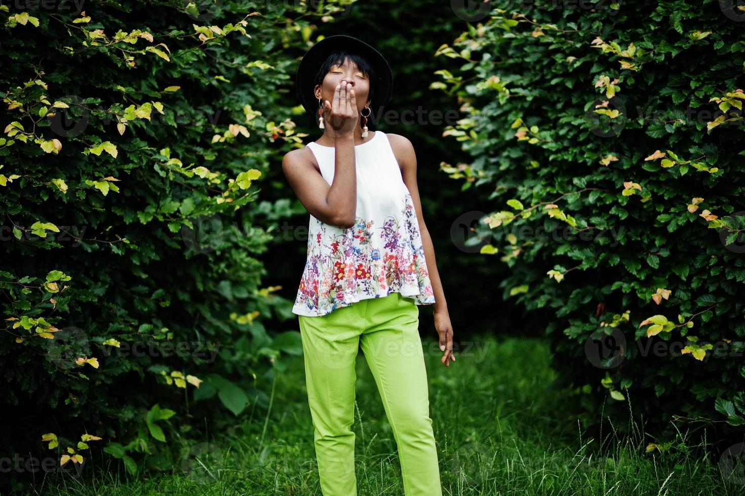 Amazing african american model woman in green pants and black hat posed with different emotions at park, shows air kiss. photo