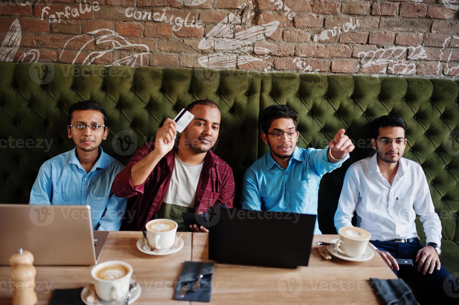 grupo de cuatro hombres del sur de asia posaron en una reunión de negocios en un café. los indios trabajan juntos con computadoras portátiles usando varios dispositivos, conversando. mantenga la tarjeta de crédito para pagar. foto