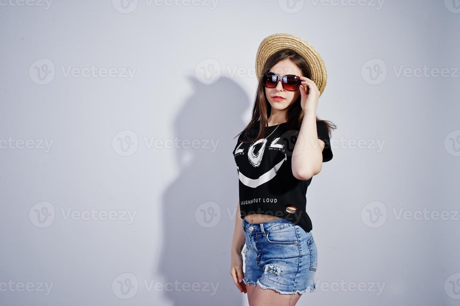 Portrait of an attractive girl in black t-shirt saying lol, denim shorts, hat and sunglasses posing in the studio. photo