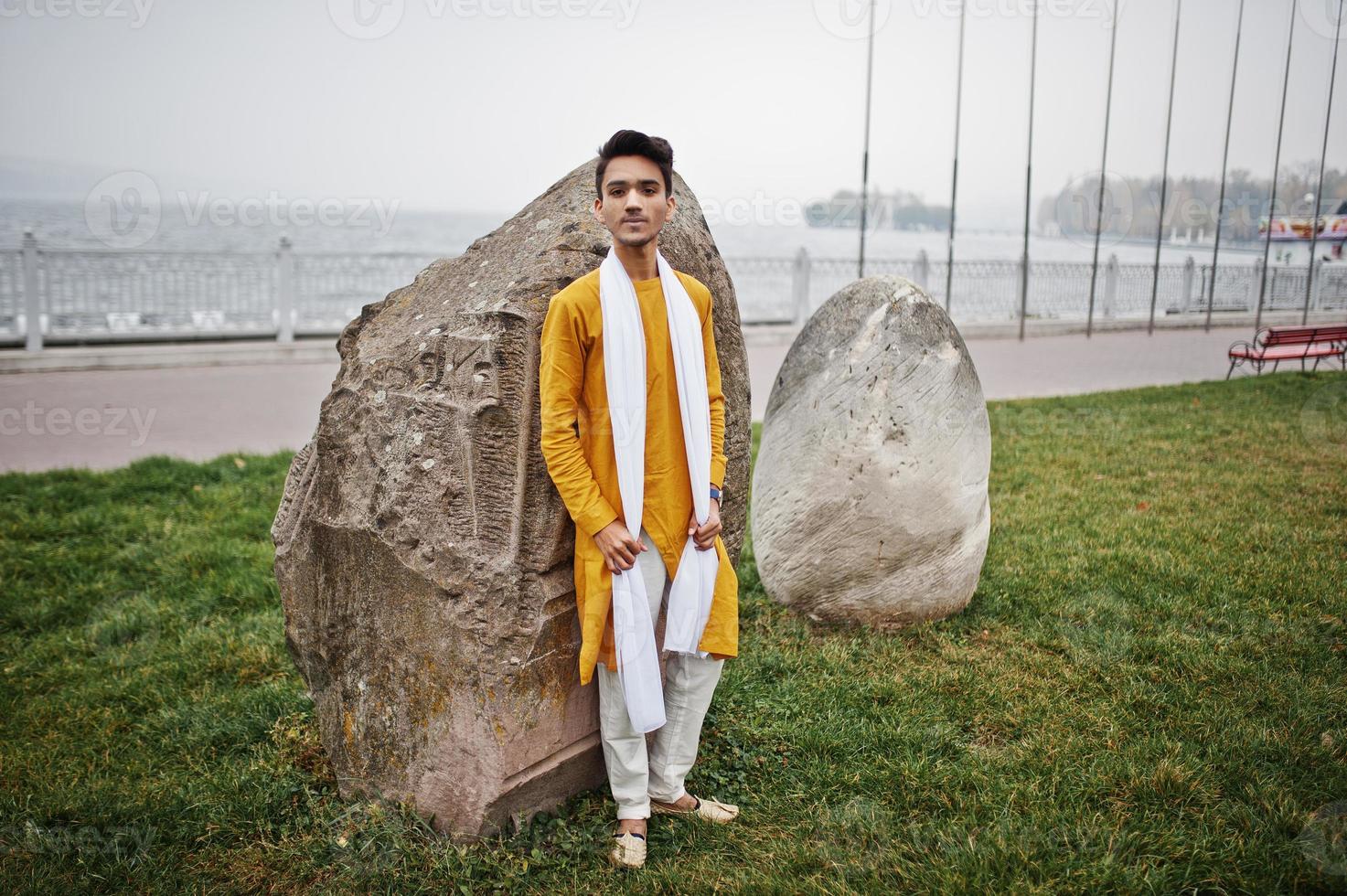 Indian stylish man in yellow traditional clothes with white scarf posed outdoor against big stones. photo