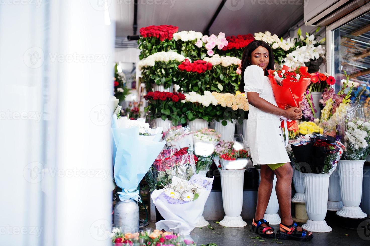 hermosa chica afroamericana en tierno vestido blanco con ramo de flores en las manos de pie contra el fondo floral en floristería.florista mujer negra. foto