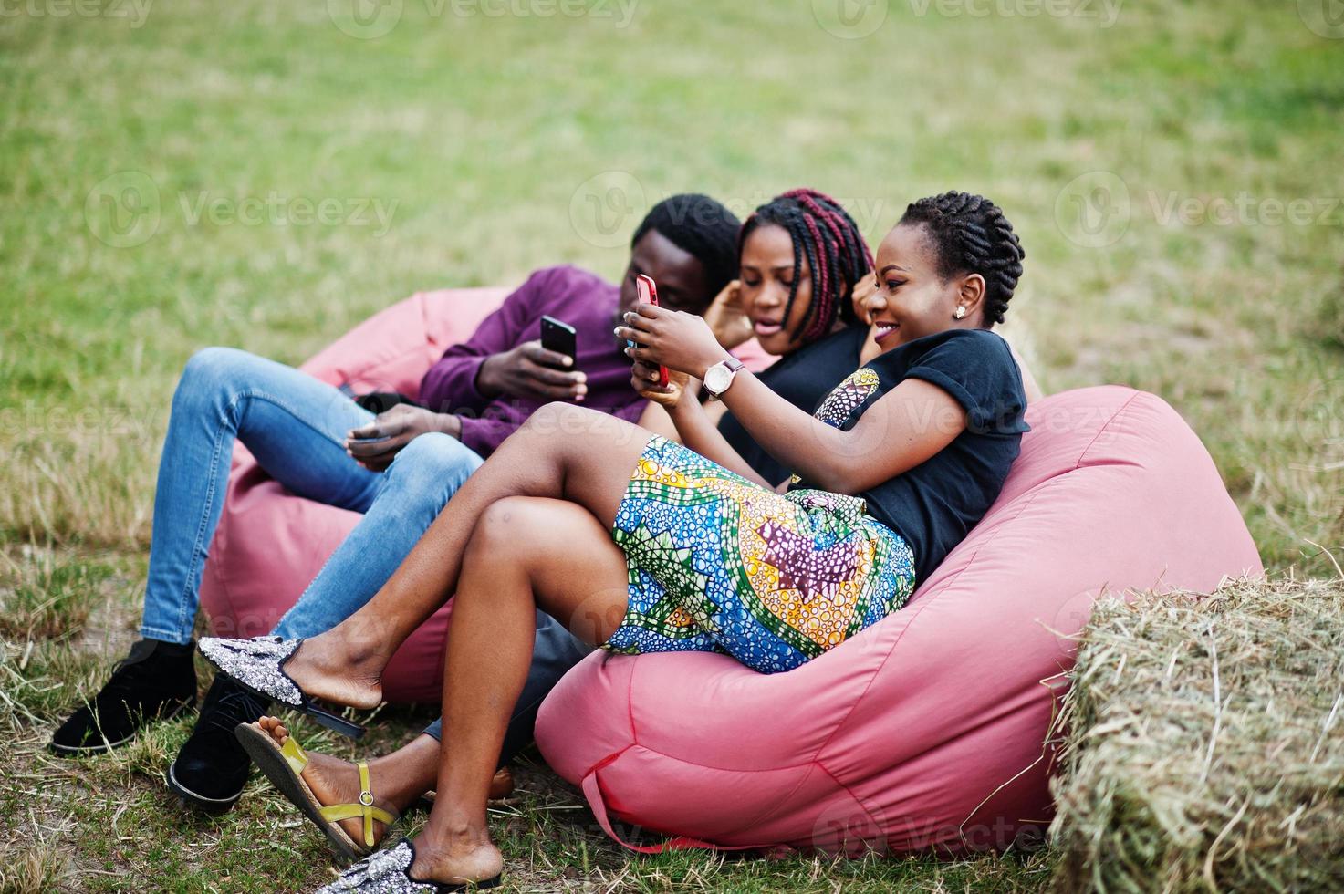 Three african american friends chill, sitting on poufs and using their phones outdoor. photo