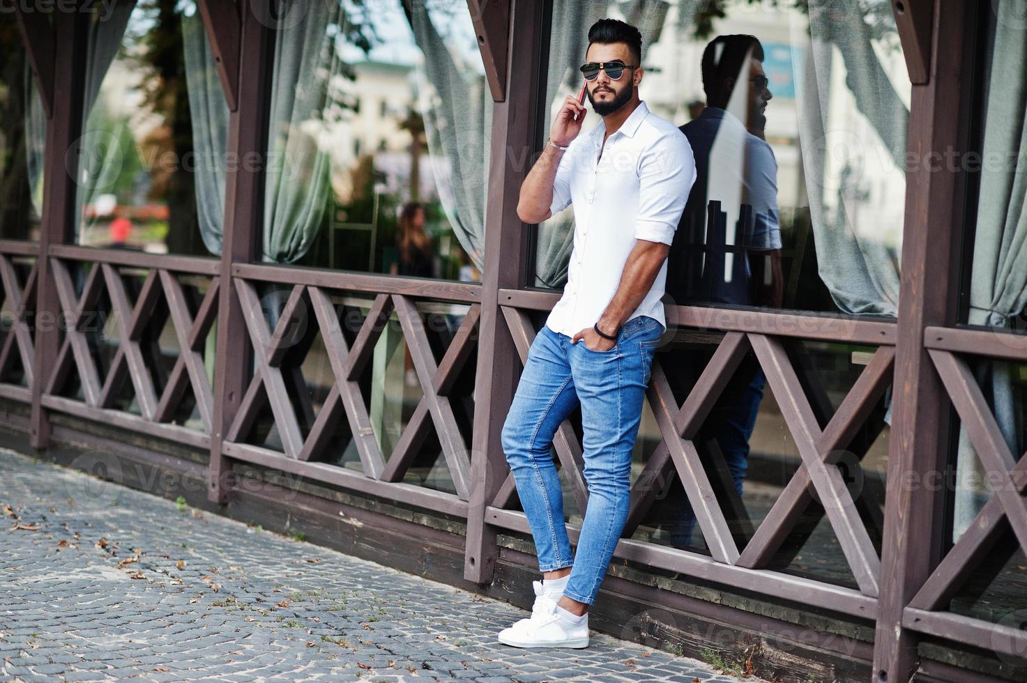 Stylish tall arabian man model in white shirt, jeans and sunglasses posed at street of city. Beard attractive arab guy speaking on mobile phone. photo