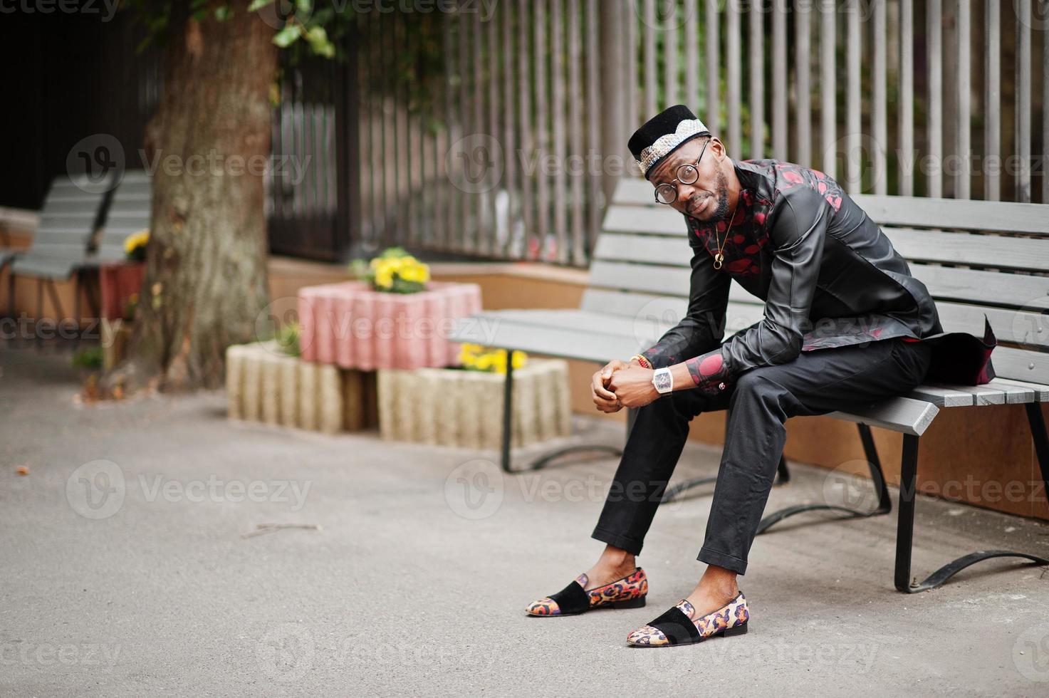 Handsome afro american man wearing traditional clothes, cap and eyeglasses in modern city sitting on bench. photo