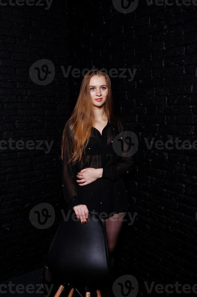 Studio shot of brunette girl in black blouse with bra and shorts against black brick wall. photo