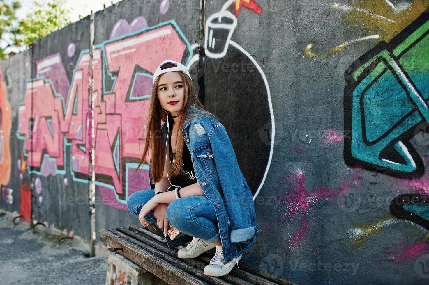 Stylish casual hipster girl in cap and jeans wear listening music from headphones of mobile phone against large graffiti wall with bomb. photo