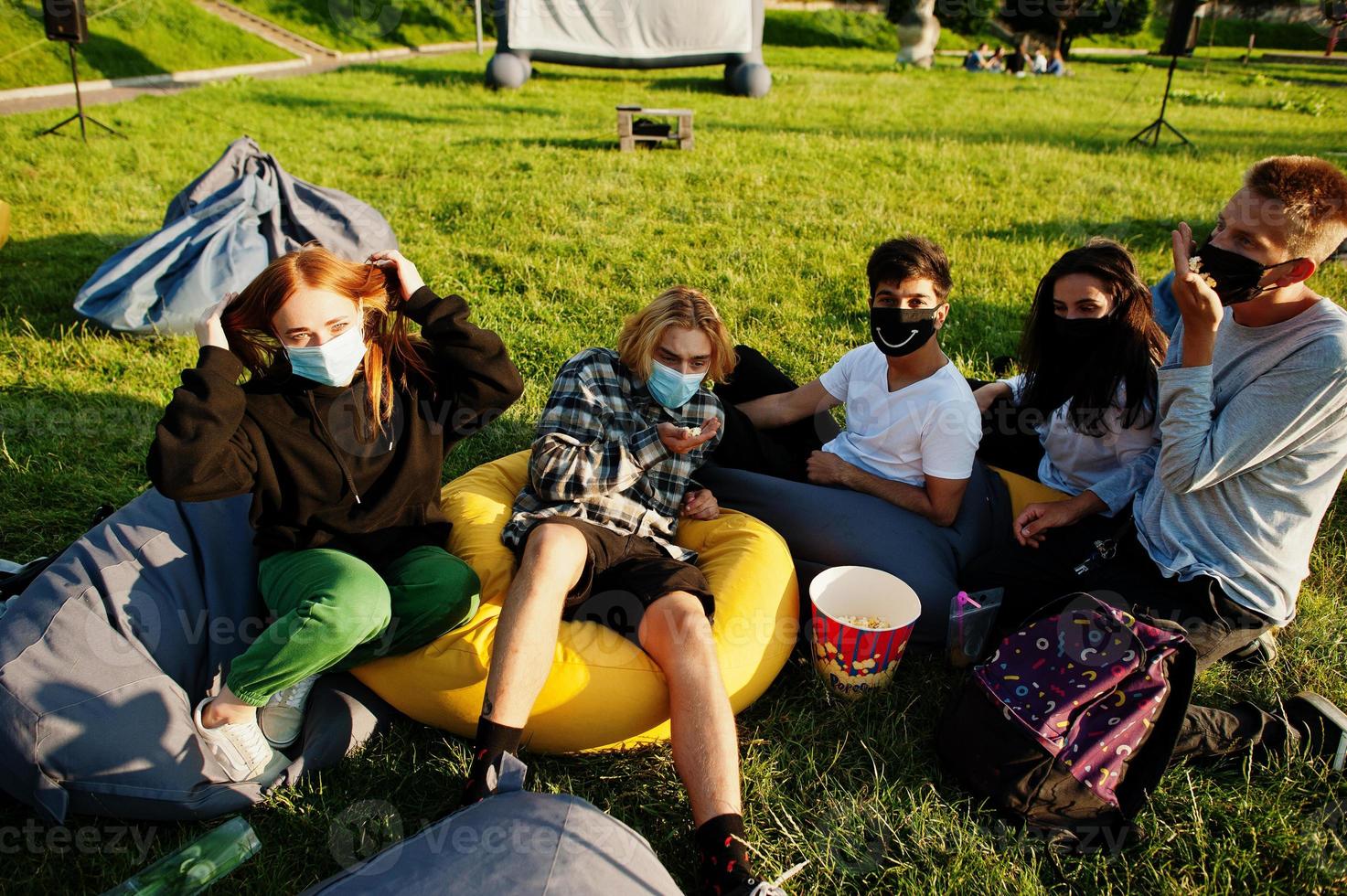 Young multi ethnic group of people watching movie at poof in open air cinema wear at mask during covid coronavirus quarantine. photo