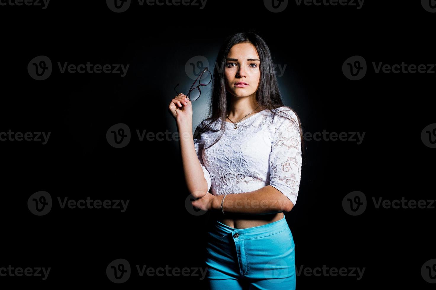 retrato de una joven atractiva con top blanco y pantalón azul posando con sus gafas en la oscuridad. foto