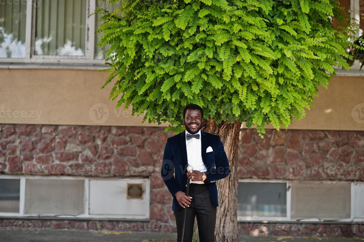 guapo hombre afroamericano de moda en ropa formal y pajarita con bastón permanecer bajo el árbol. foto