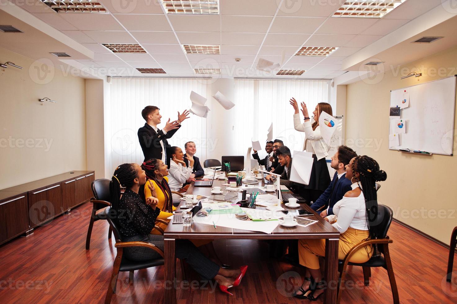 Multiracial business team meeting around boardroom table, two team leaders throw paper up. photo