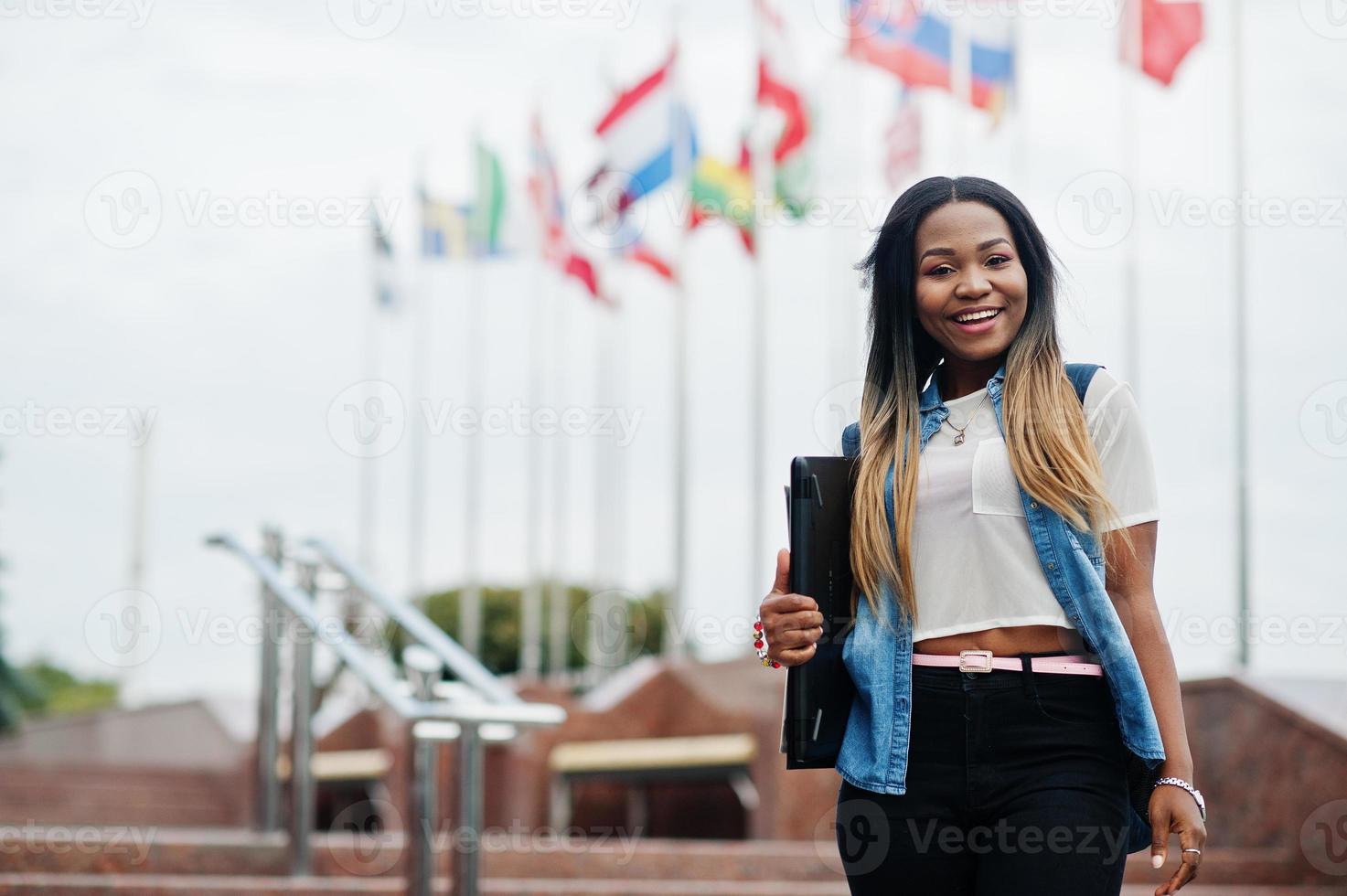 African student female posed with backpack and school items on yard of university, against flags of different countries. photo