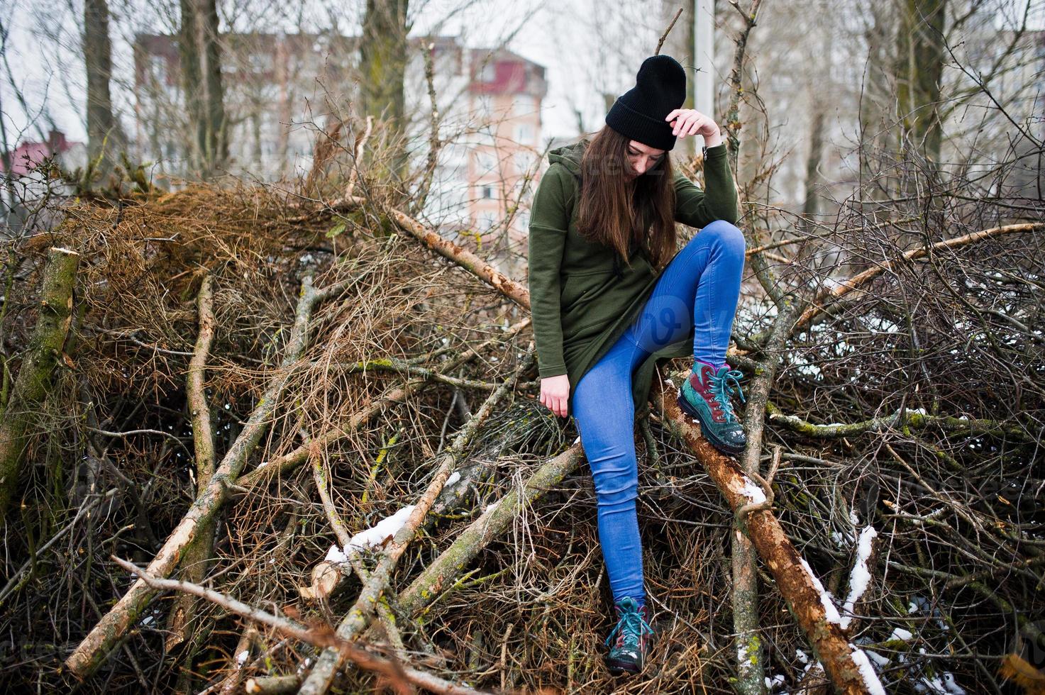 la niña usa una sudadera verde larga, jeans y sombreros negros en las ramas del pino en el día de invierno. foto