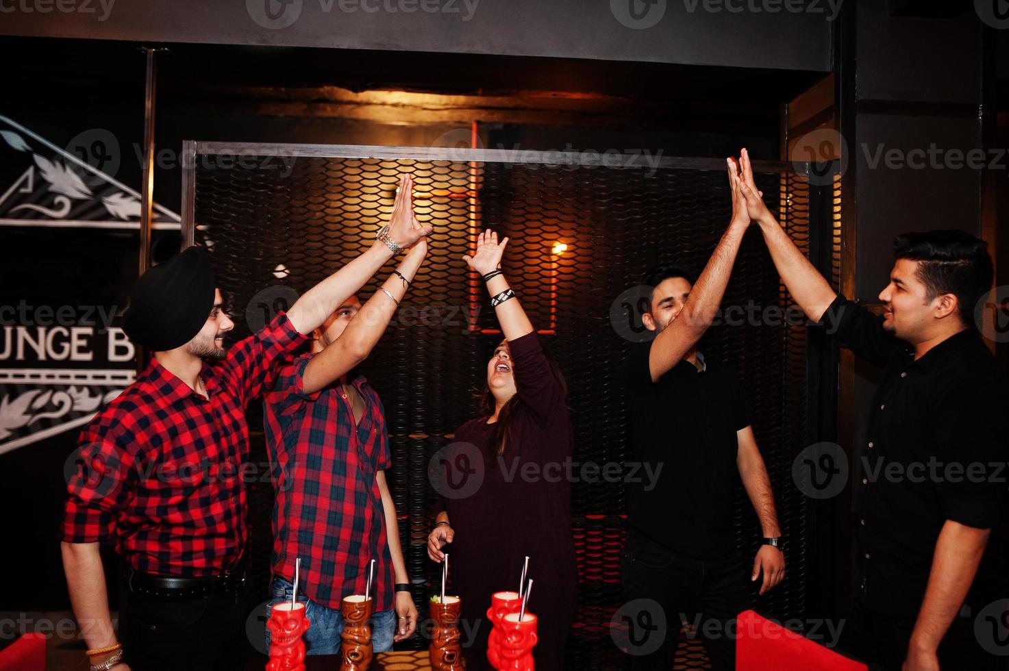 Group of indian friends having fun and rest at night club, drinking cocktails and giving high five together. photo