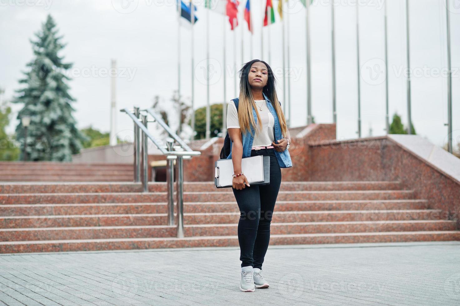 African student female posed with backpack and school items on yard of university, against flags of different countries. photo