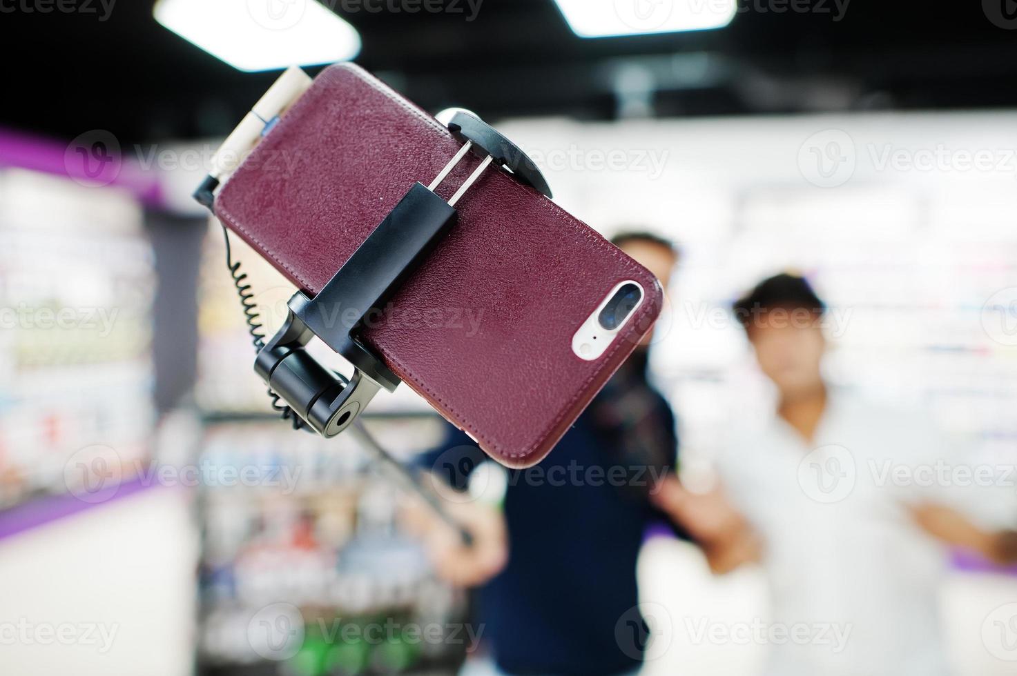 Close up photo of mobile phone on monopod stick against two indians mans customer buyer aking selfie. South asian peoples and technologies concept. Cellphone shop.