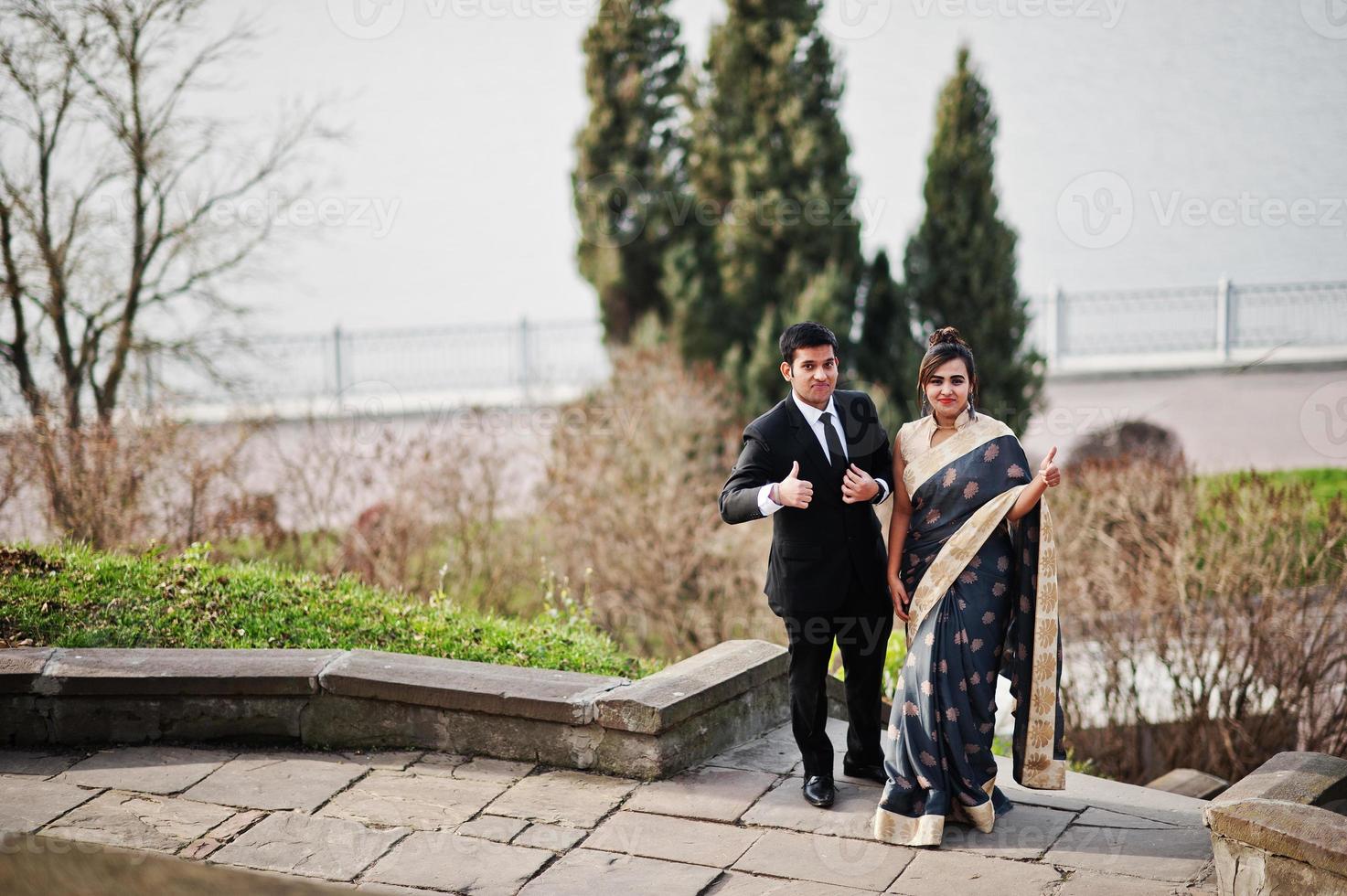 Elegant and fashionable indian friends couple of woman in saree and man in suit posed on stairs against lake. photo