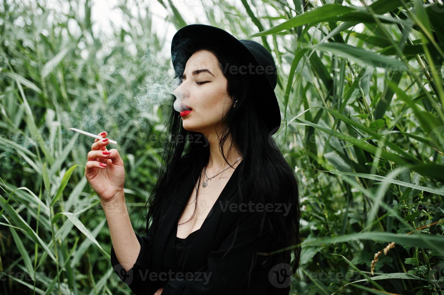 Sensual smoker girl all in black, red lips and hat. Goth dramatic woman smoking on common reed. photo