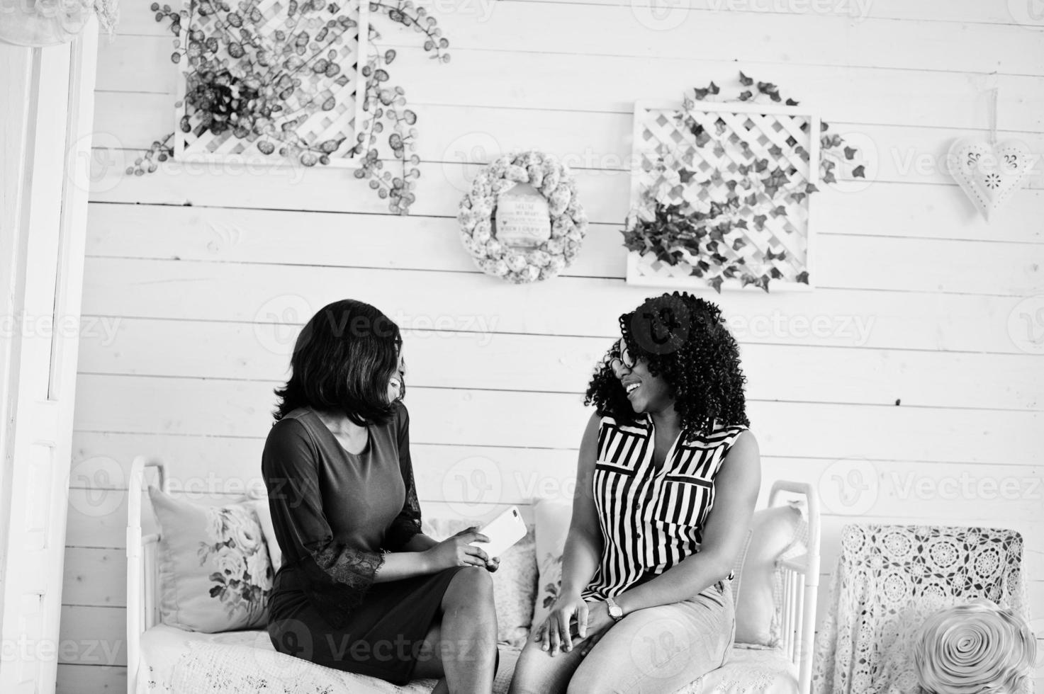 Two african woman friends wear on eyeglasses sitting at couch indoor white room. They chatting, one of them hold mobile phone. photo