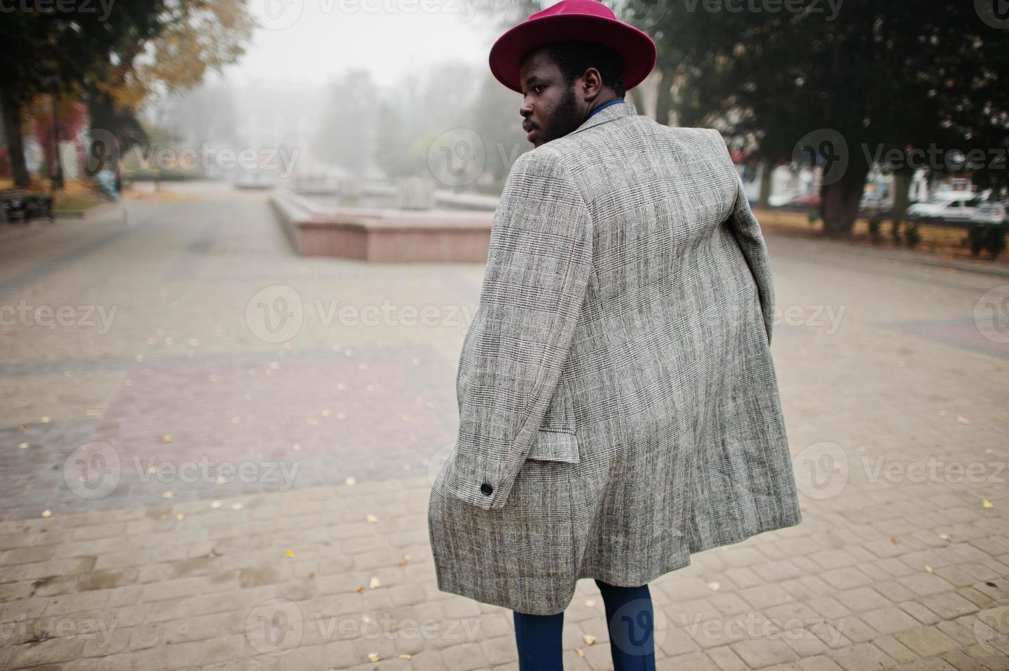 elegante modelo afroamericano con abrigo gris, corbata de chaqueta y sombrero rojo caminando en la calle con niebla. foto