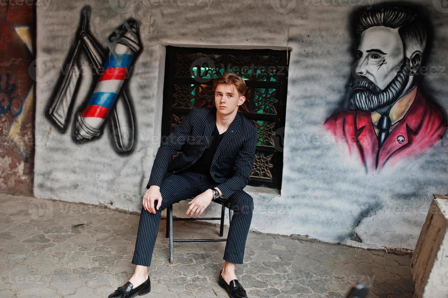 joven macho con estilo en chaqueta negra planteada al aire libre de la calle. asombroso modelo de hombre sentado contra la pared de grafiti de la barbería. foto