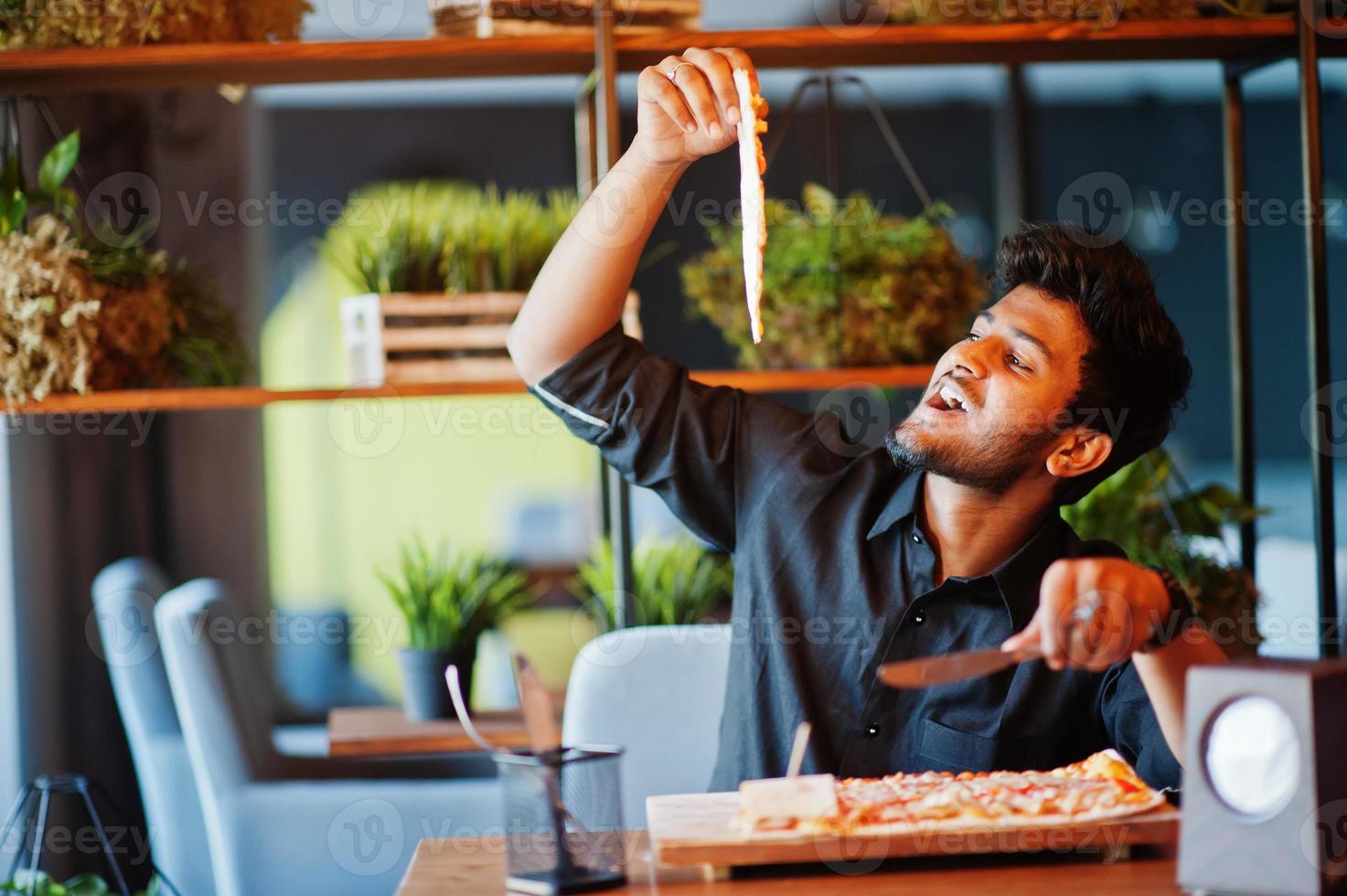 un joven indio confiado con camisa negra sentado en una pizzería con pizza. foto