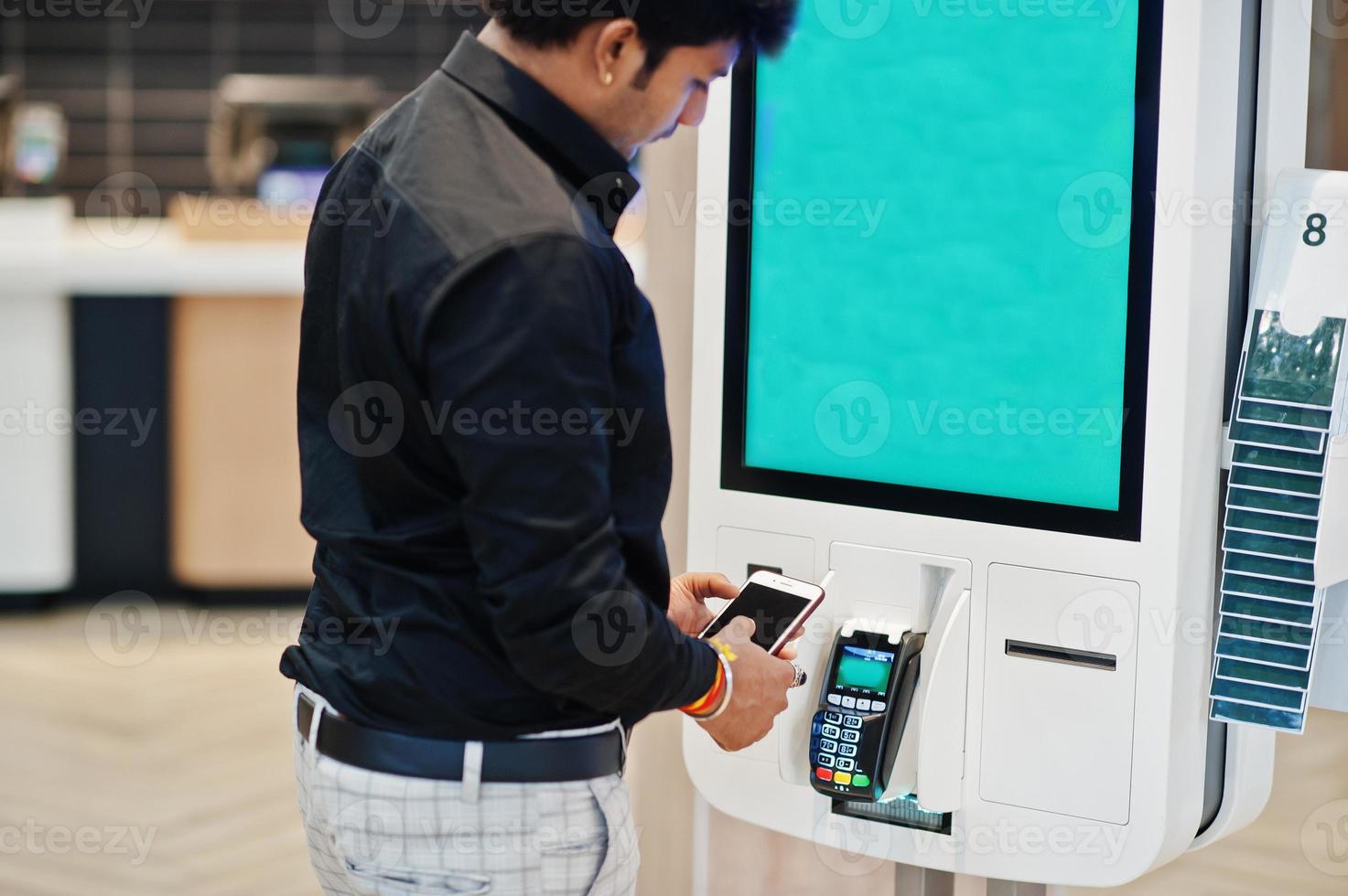 Indian man customer at store place orders and pay by contactless credit card on mobile phone through self pay floor kiosk for fast food, payment terminal. Pay pass. photo
