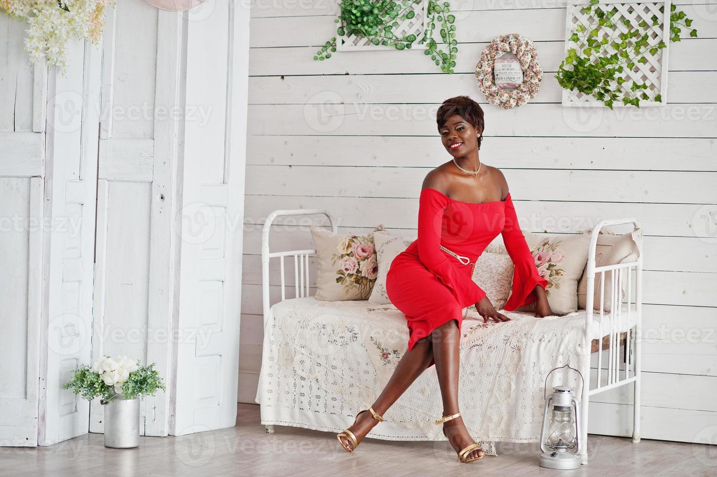 Fashion african american model in red beauty dress, sexy woman posing evening gown sitting at white vintage room. photo