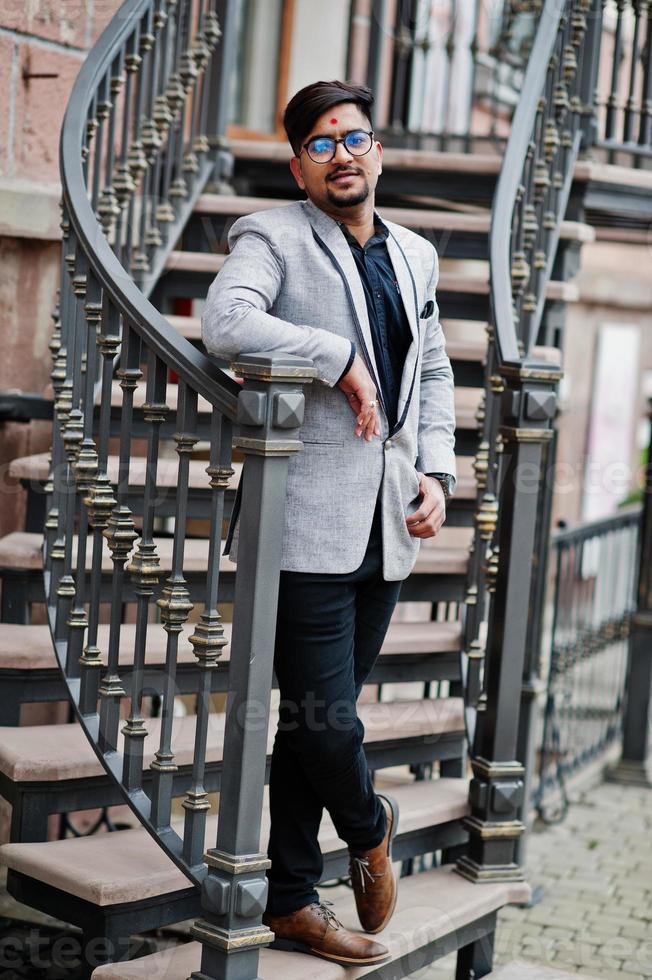 Stylish indian man with bindi on forehead and glasses, wear on suit posed outdoor against iron stairs. photo