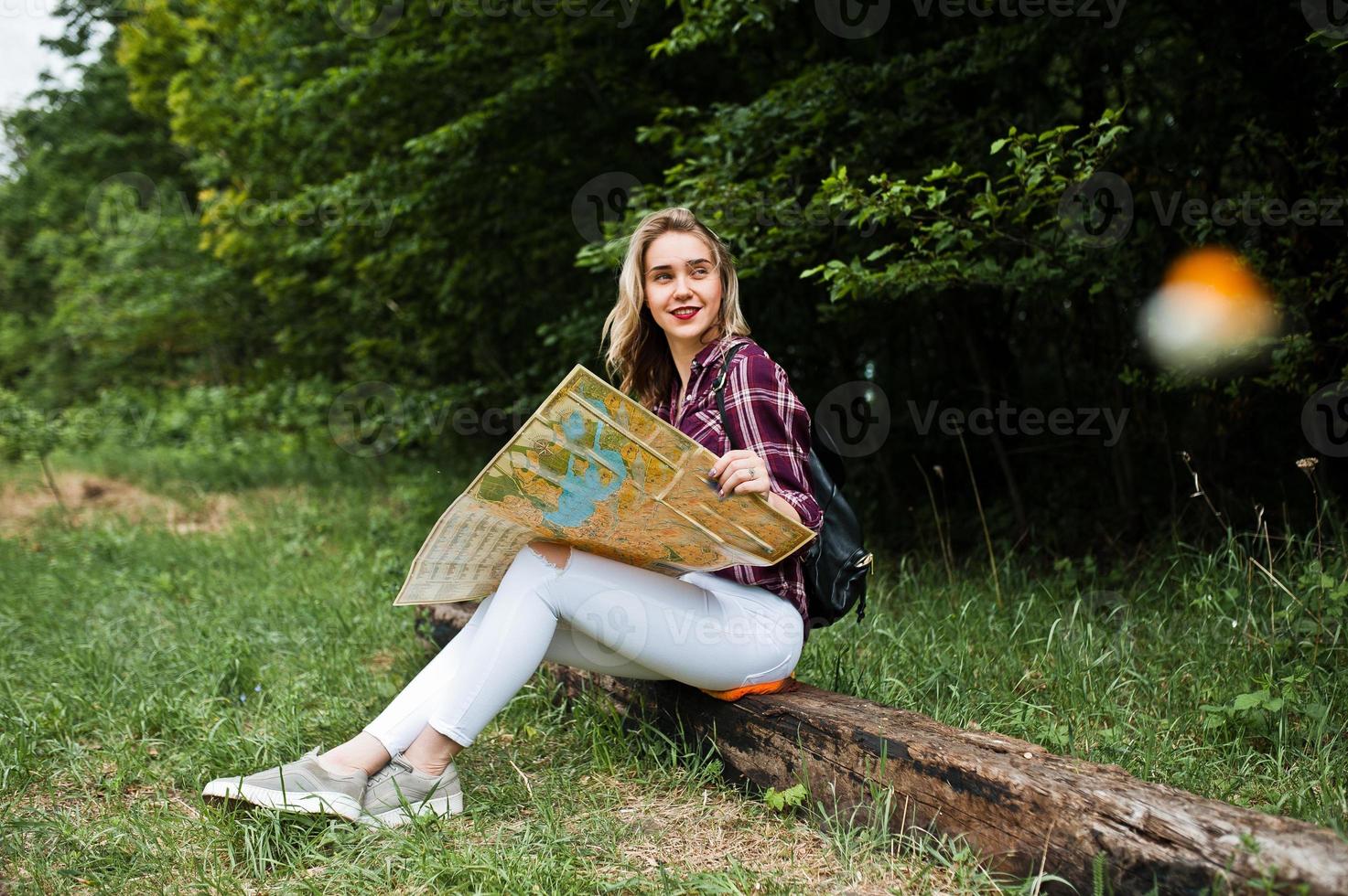 Portrait of a positive young gorgeous blonde sitting on the ground with a map in her hands in the forest. photo