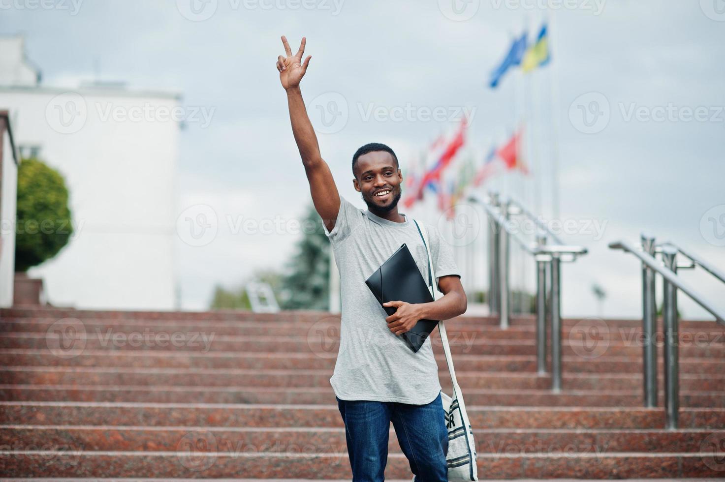 African student male posed with backpack and school items on yard of university, against flags of different countries. Show two fingers. photo