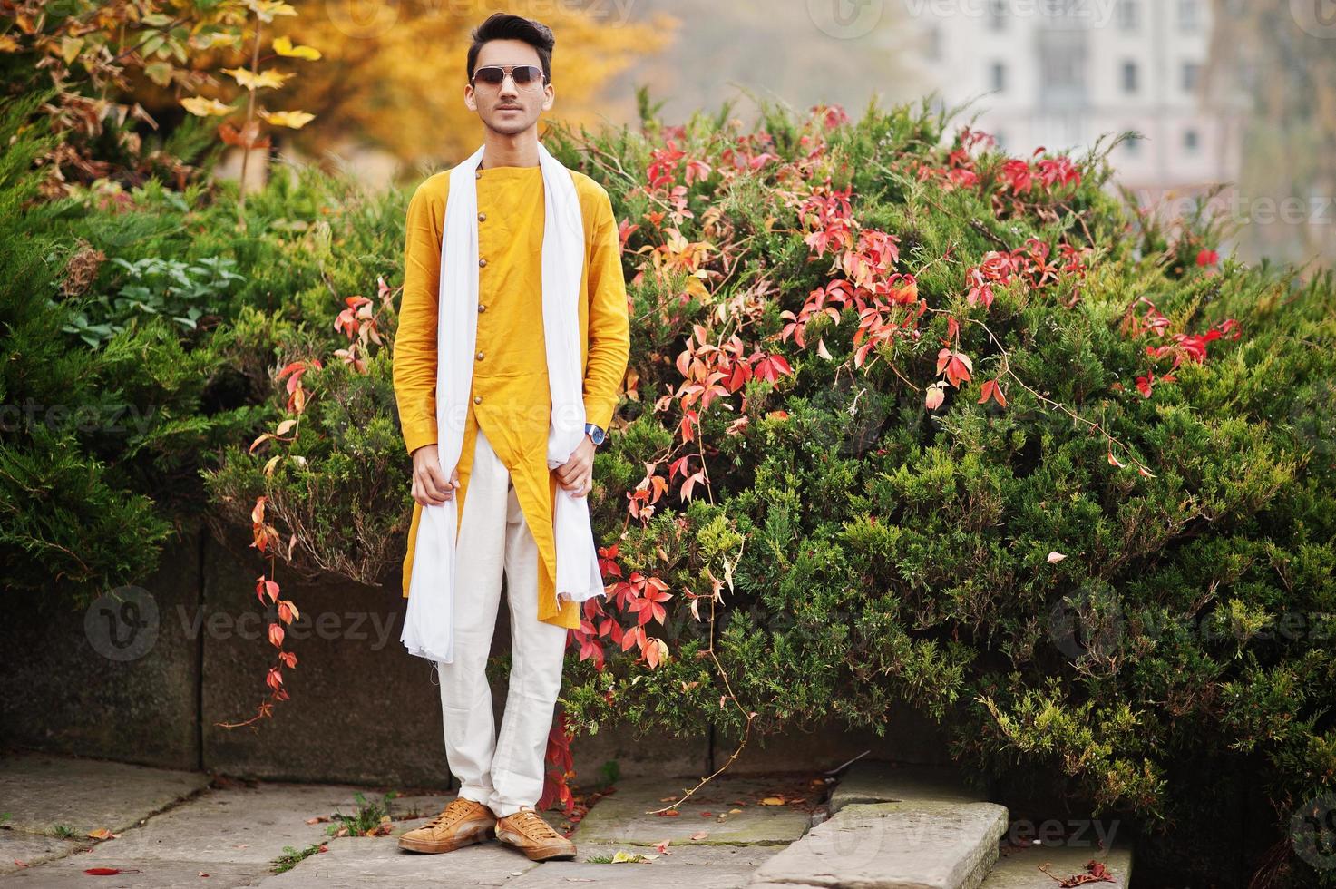 Indian stylish man in yellow traditional clothes with white scarf, sunglasses posed outdoor against autumn leaves tree. photo